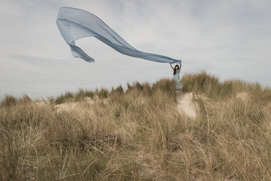 Girl in a blue dress standing on dune holding and flying a large blue thin drape in the wind