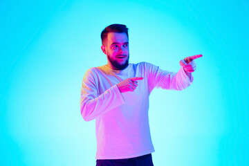 Naklejka premium Pointing, choosing. Caucasian man's portrait isolated on blue studio background in neon light. Beautiful male model. Concept of human emotions, facial expression, sales, ad. Copyspace for ad.
