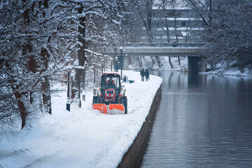 Tractor with snow plow and rotating brush sweeping snow from foopath on embankment in park. Red...