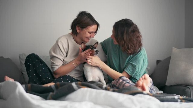 Beautiful lesbian couple cuddling their cute basenji dog while sitting on the bed in pajamas. Cozy moments with a pet concept.