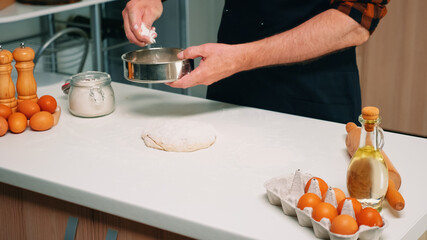 Senior man sifting flour with metallic sieve over dough. Happy elderly chef with kitchen uniform mixing, sprinkling, adding, sifting spreading raw ingredients to baking traditional bread