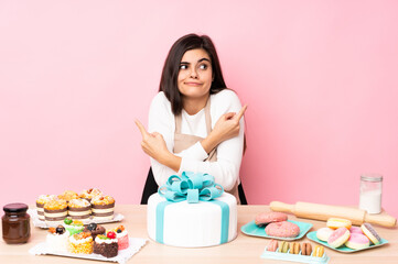 Pastry chef with a big cake in a table over isolated pink background pointing to the laterals having doubts