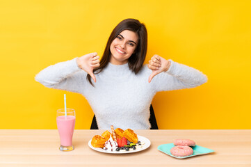 Young woman eating waffles and milkshake in a table over isolated yellow background proud and self-satisfied