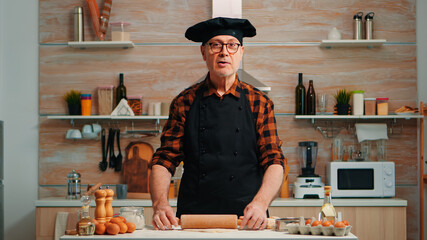 Elderly man rolling dough at home infront of video camera explaining recipe step by step. Retired blogger chef influencer using internet technology communicating on social media with digital equipment