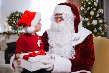 Santa Claus giving present to little boy in room decorated for Christmas