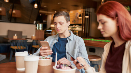 Portrait of couple of teenagers totally absorbed in using their phones, ignoring each other while sitting in a cafe together on a daytime