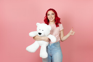 a smiling, delighted young woman with red hair holds a large white teddy bear and gives a thumbs up isolated on a pink background.