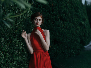 Fototapeta premium Beautiful woman woman in red dress outdoors near bush luxury charm