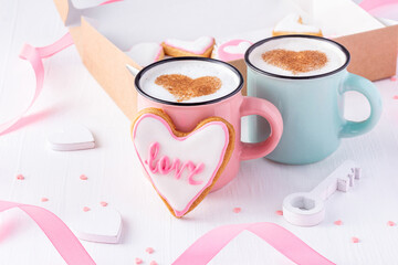 A couple of mugs with latte decorated with cinnamon hearts nearby heart shaped cookies glazed with word love on white wooden background for St Valentine day