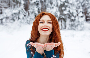 Young red-haired girl with long red hair on a winter background.Air kiss. Red-haired woman in a summer dress on the background of winter nature. - Powered by Adobe