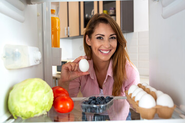 Smiling beautiful woman holding an egg seeming eager to prepare some food with the groceries from the refrigerator