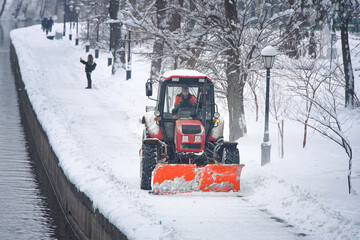 Snow removal tractor with rotating brush sweeping snow from footpath on embankment in park. Red tractor with scoop and automated brush removes snow. Municipal road sweeping vehicle with plow