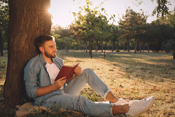 Fototapeta na wymiar Young man reading book on green grass near tree in park