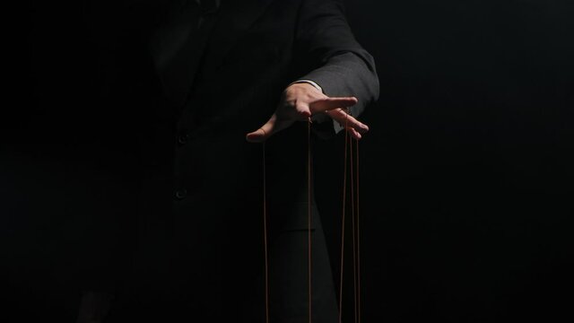 Puppet Master controls and manipulates the puppets with strings attached to his fingers. Hand of businessman in a suit close up controls destinies of people. Isolated on black background. Slow motion.