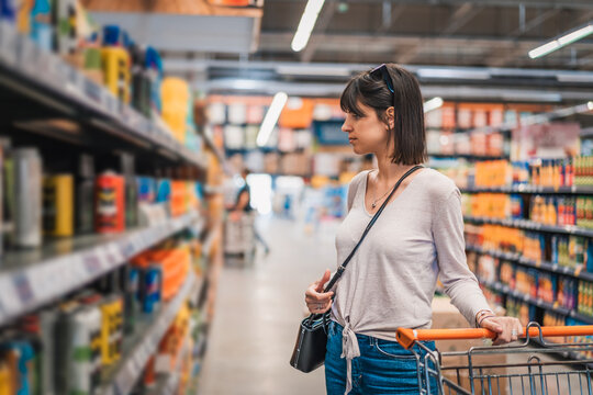 Sale, shopping, consumerism and people concept. Woman in supermarket with shopping trolley choosing products and looking aside. Female chooses products on the background of shelvesin a supermarket. 