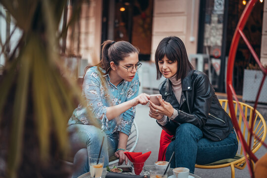 Two beautiful women in cafe outdoors looking at something on mobile phone. Two female using mobile phone application shopping online and enjoying coffee outside in city