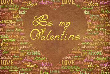yellow ink inscription "be my valentine" on a cardboard heart against a background of multicolored handwritten words "love" in english and all european languages