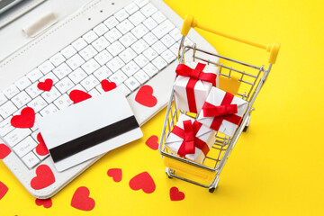 Shopping online during holidays. Ordering Valentines day gifts. Shopping cart with gift boxes, credit card, hearts and white laptop on yellow background.