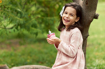 Image of a cheerful little girl smiles and makes a wish with the birthday cupcake in her hands in...