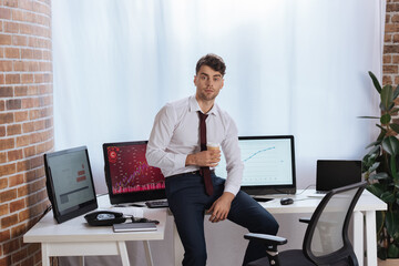 Forex businessman with coffee to go looking at camera near computers and telephone on table