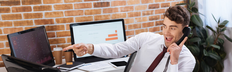 Cheerful businessman talking on telephone and pointing at computer while checking forex courses, banner