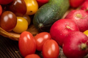 Green cucumbers, ripe radishes and colorful tomatoes in a wicker basket and scattered on the table. Close-up, selective focus.