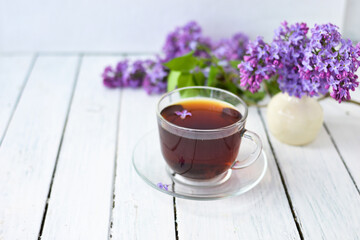 Delicate setting of the morning tea table with lilac flowers, a transparent cup of tea and a saucer and a white vase on a white wooden board. Copy space. Spring breakfast concept