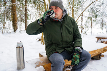 a guy in a winter forest drinks a hot drink from a thermos bottle, a tourism concept - obrazy, fototapety, plakaty