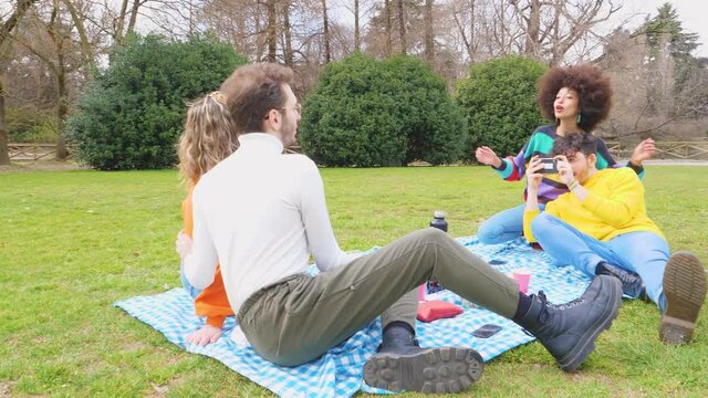 Four cheerful friends diverse multiethnic having fun doing pic nic in a park outdoors using smartphone taking photos making memories enjoying quality time together