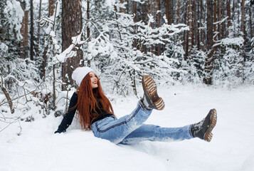 Fototapeta na wymiar A happy girl with bright red hair is sitting on the snow in a snow-covered forest.