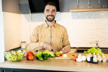 Handsome guy with beard and nice smile is cooking salad. He probably likes healthy lifestyle and eating fresh food. Young vegetarian man is cooking in kitchen indoor