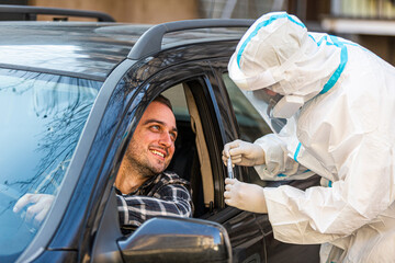 Man sitting in car, waiting for medical worker in PPE to perform drive-thru COVID-19 test