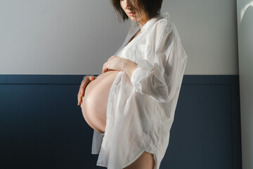 Young pregnant girl in white underwear and white shirt near a white wall in the sun