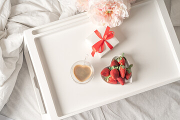Breakfast for Valentine's Day. Heart shaped white plate with fresh strawberries, cup of coffee and flowers with gift in bed. Still life composition. Flat lay, top view, Mother's Day