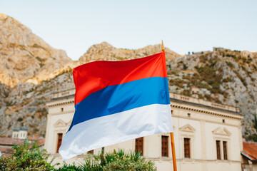 The flag of Serbia flies against the backdrop of a building and mountains on a sunny day.