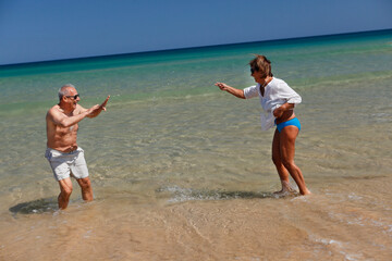 Fototapeta na wymiar Coppia di anziani , gioca e si diverte sulla riva di una spiaggia in abbigliamento estivo