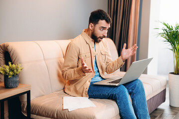 Programmer working from home is shocked reading work chat. Young bearded man in casual clothes sits on smart modern sofa with laptop on his knees, his hands up in astonished gesture. Remote work.