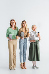 Fototapeta premium full length of three generation of cheerful women holding newspapers and paper cups on white