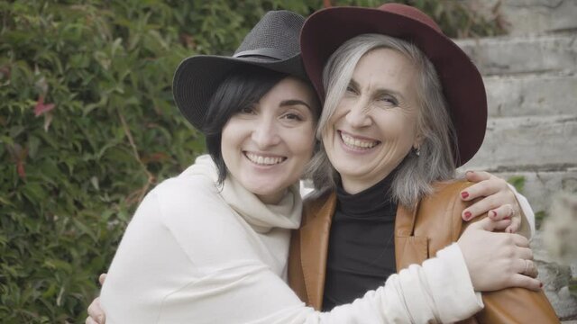 Loving adult daughter adjusting elegant hat on head of gorgeous senior mother sitting in park. Portrait of happy Caucasian women looking at camera and smiling resting outdoors. Family concept.