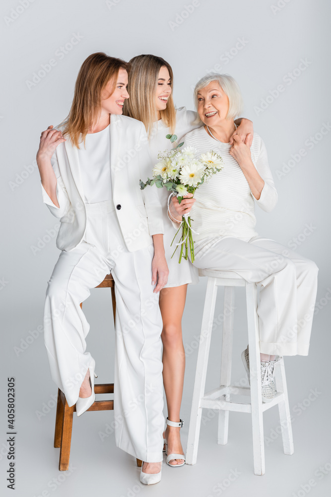 Sticker full length of three generation of happy women smiling while hugging near flowers on grey