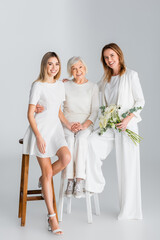 full length of happy woman holding flowers and smiling while sitting on chair with daughter and...
