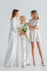 full length of happy senior woman holding flowers and smiling with daughter and granddaughter on grey