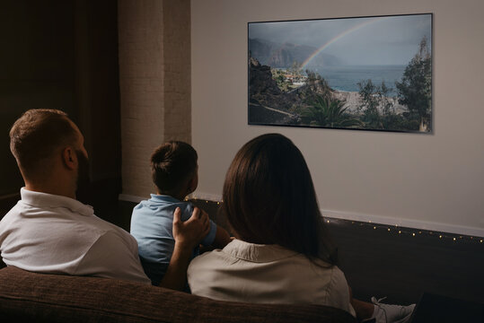 A Photo From Behind Of A Father, A Son, And A Young Mother Which Are Watching A Movie On A Widescreen Television Set On The Sofa. The Family Is Enjoying A Video Together In The Evening At Home.