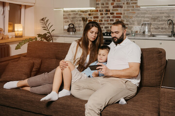 Obraz premium A dad, a son, and a young mother are watching a video on a smartphone on the sofa. A father with a beard is demonstrating to his family news on the screen of the cellphone in the evening at home.