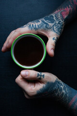 Tattooed man's hands holding a cup of coffee on dark background