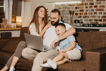 A father is trying to work remotely on a laptop near his smiling son and a curious wife at home. A family at the sofa in the evening. A dad is working online on a computer between relatives.