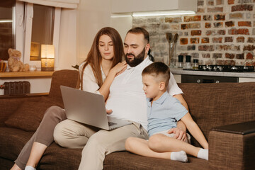 A serious father with a beard is trying to work remotely on a laptop near his son and a curious wife at home. A family at the sofa in the evening. Dad is working online on a computer between relatives