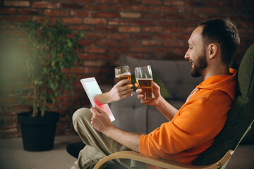 Clinking. Young man drinking beer during meeting friends on virtual video call. Distance online meeting, chat together on tablet at home. Concept of remote safe meetings and entertainment.