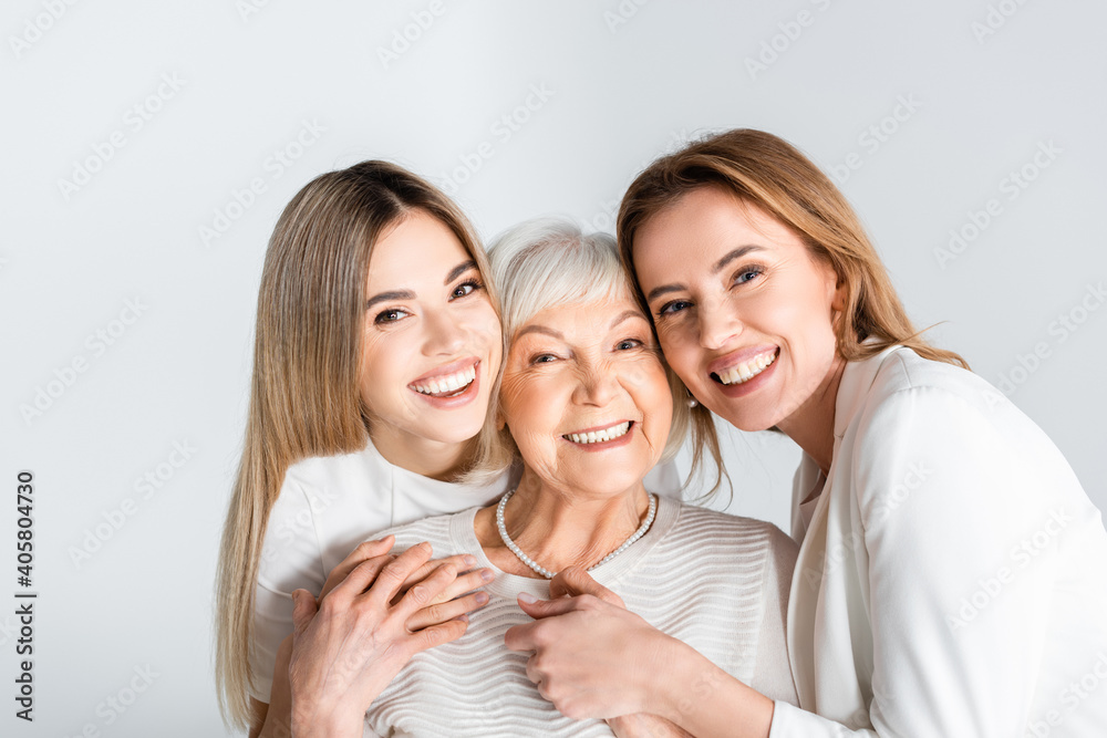 Wall mural three generation of positive women smiling while looking at camera and hugging isolated on grey