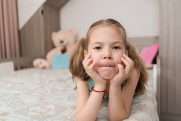 Lovely carefree kid portrait making sweet, funny faces while lying on her bed in her room
